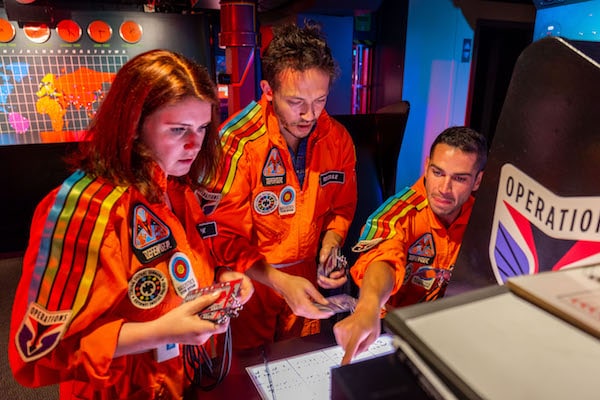 In-game: 3 players in orange flightsuits working at the Operations cabinet.