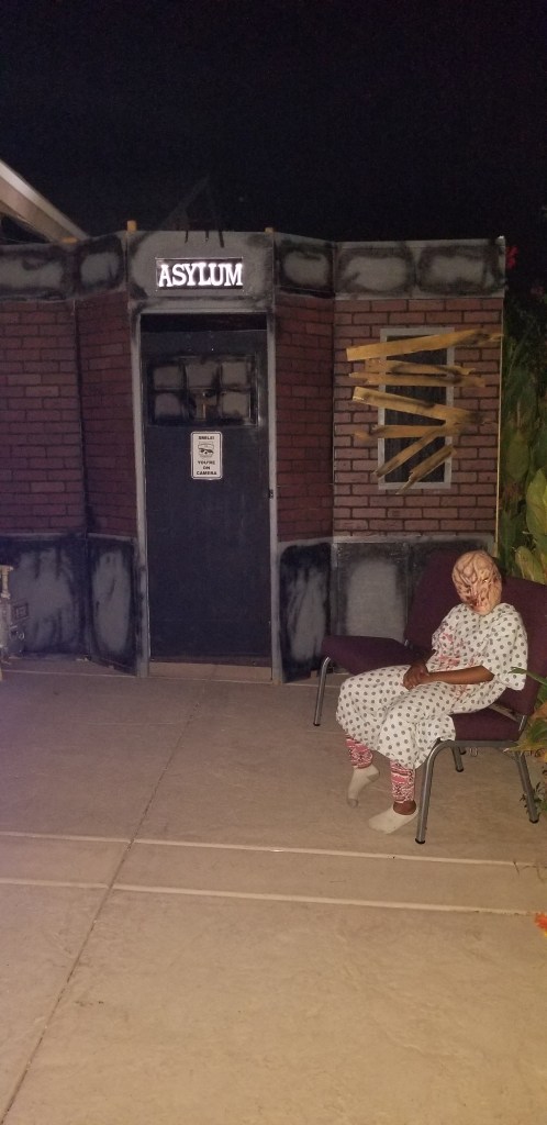 facade of haunt wall titled asylum, patient sitting with scary mask