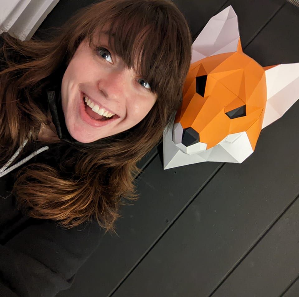smiling woman with bangs and long brunette hair in a black sweatshirt posing with a paper fox mask