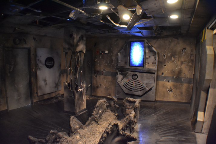 photo of a room designed to look like a bomb exploded. the column is broken with exposed wire. The ceiling is collapsing and the center table is blown up.