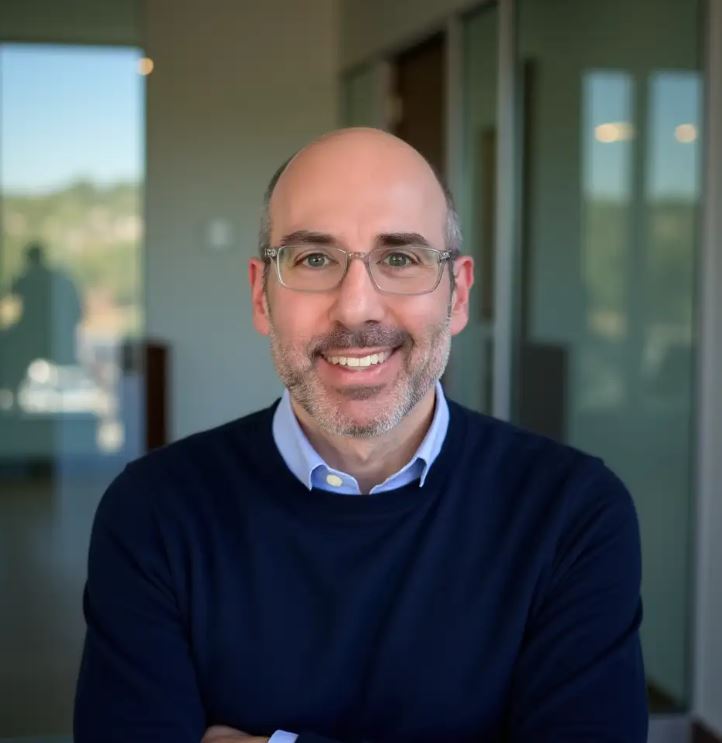 man with horseshoe hairline, wearing glasses and a short salt and pepper beard, smiling, wearing a blue sweater over a light blue button down shirt.