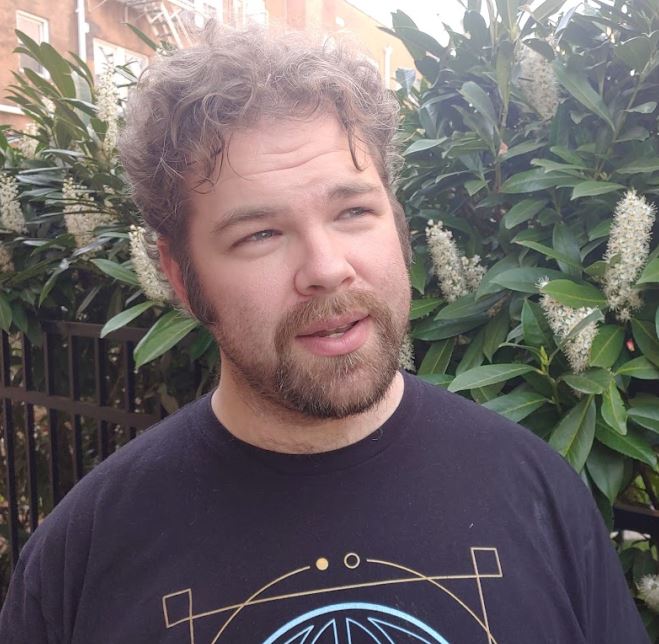 man with curly brown hair and short beard, wearing a black t-shirt and gazing to the side.