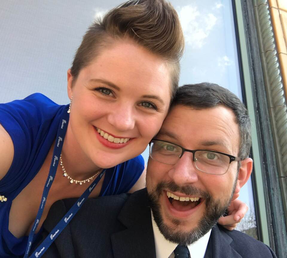 woman with mohawk smiling in a blue dress with a man in glasses and beard, with short brown hair wearing a suit