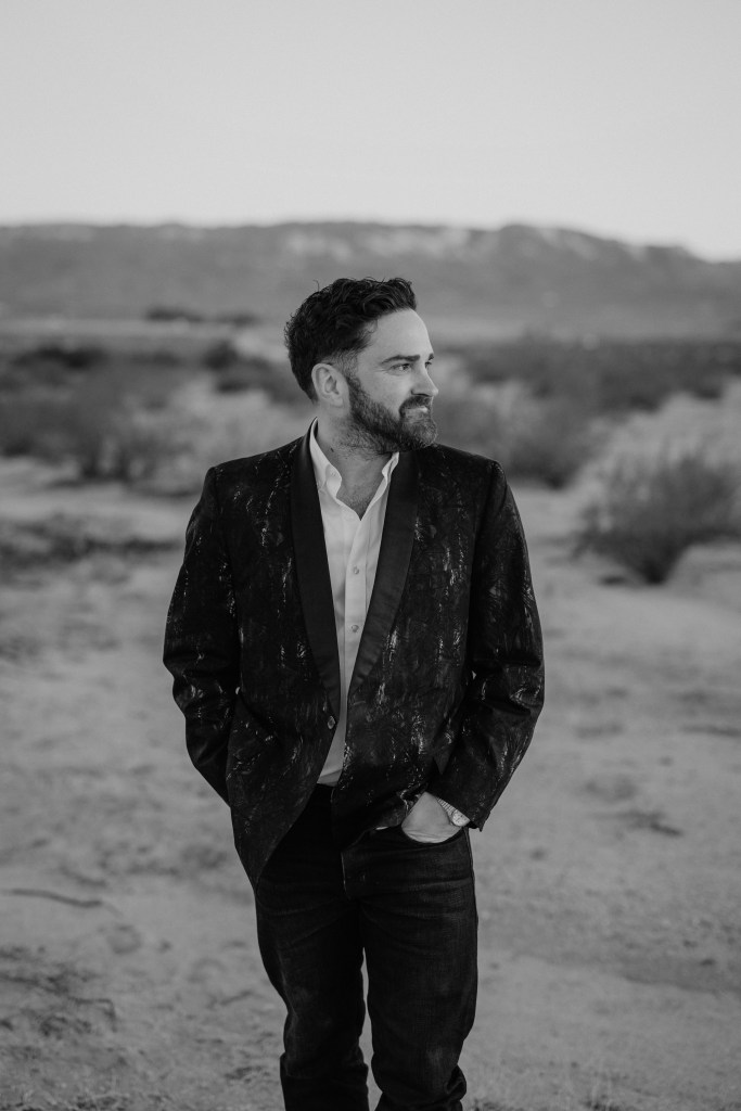 black and white image of man in suit and black jeans in desert looking to the right. short wavy hair and short beard.