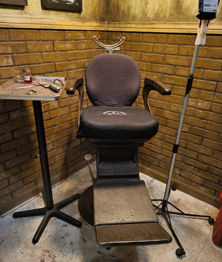 A beat up, old, dentists chair beside a tray with medical equipment and a bag of blood on a pole.