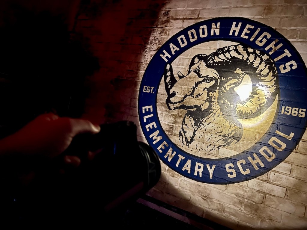 A hand holding a flashlight to a the RAM logo of Haddon Heights Elementary School painted on a brick wall.