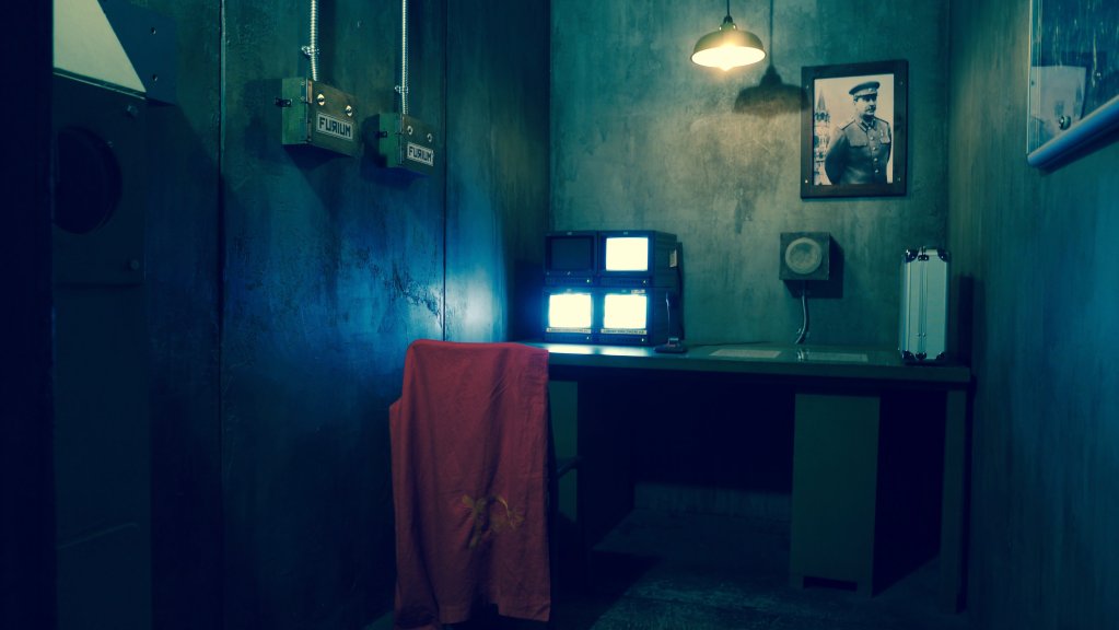 room with concrete walls and a photo of a military man on the wall. There is a desk with monitors and equipment.