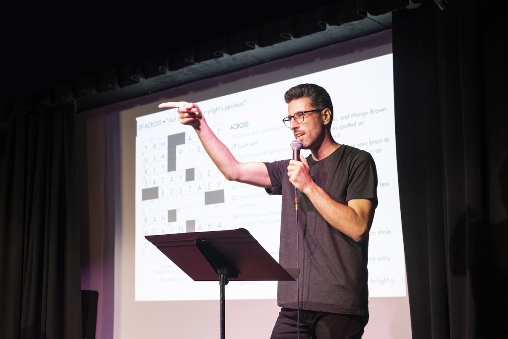 Zach Sherwin with a microphone on stage with a crossword projected behind him.