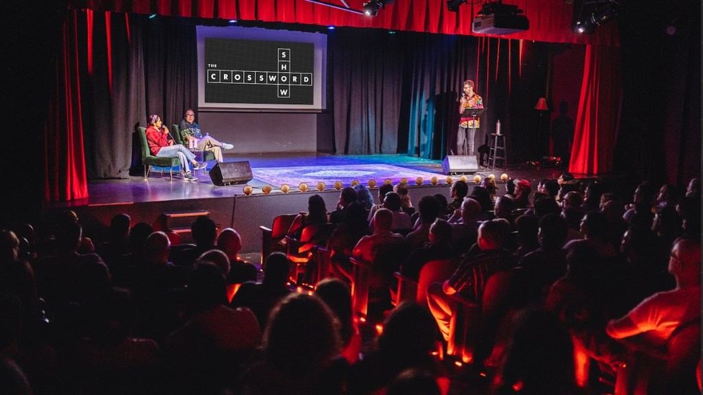 Zach Sherwin with a microphone on stage bantering with guests, the Crossword Show logo is projected behind them.