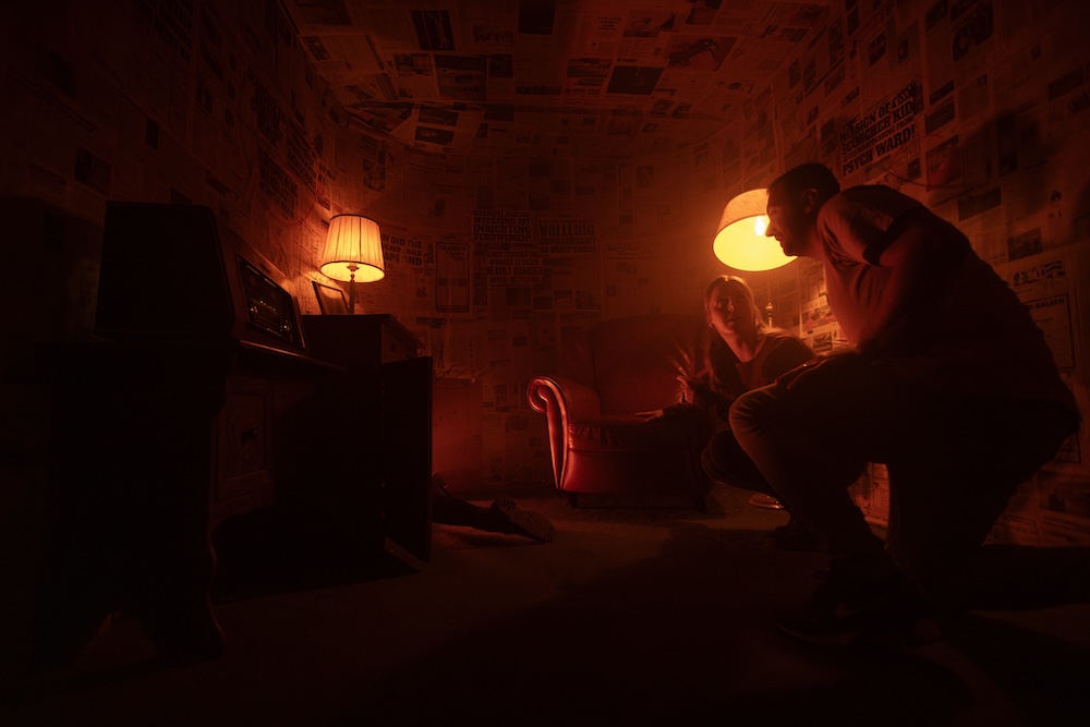 Two people kneel in a strange living room that is wallpapered in newsprint.