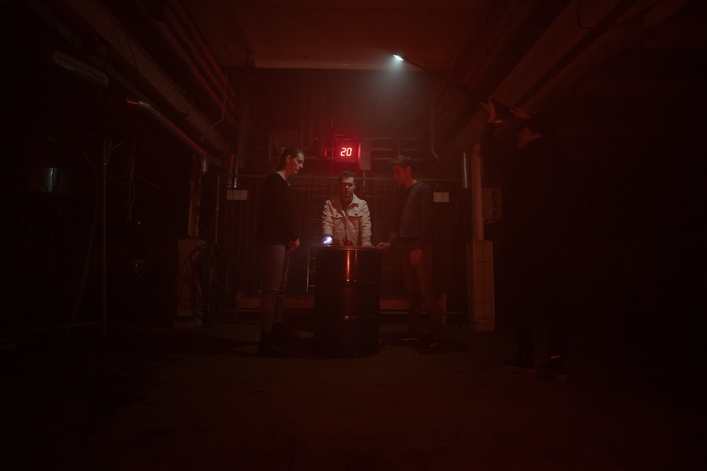 3 people stand around a pedestal in a gloomy factory. A red LED 20 illuminates behind them.