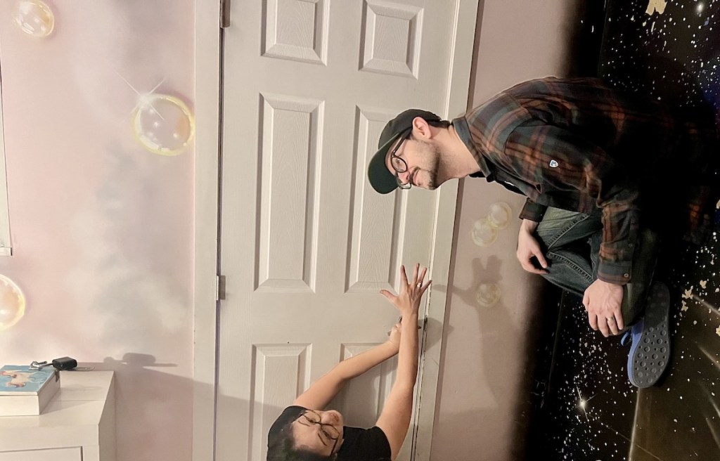 Impossible of a man sitting sideways looking downward in front of a vertical door, while a woman climbs upwards reaching out towards him.