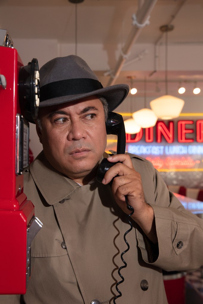 filipino man wearing a trench coat and fedora, clean shaven, holding a telephone receiver from a red public payphone to his ear. Background is a neon sign that says "diner"