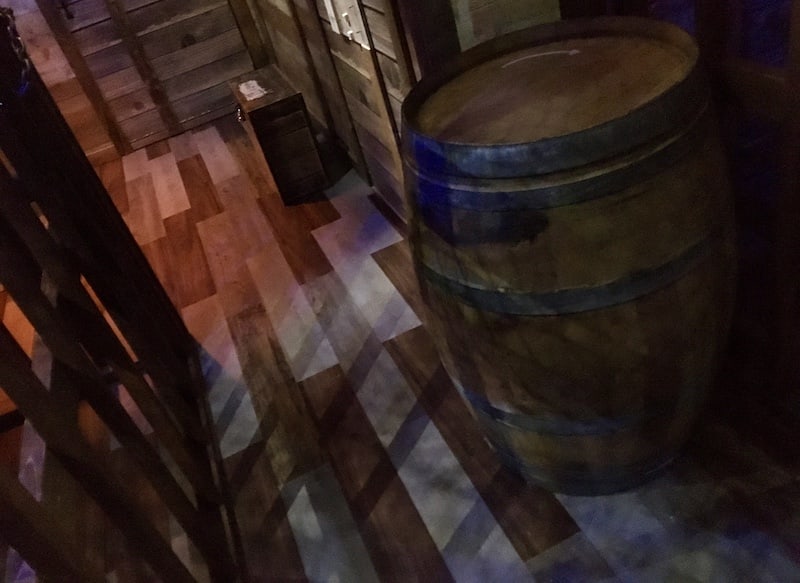 In-game: The wooden interior of a ship, a large barrel in the middle of the shot.