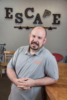 Man with short hair and short brown beard leaning on a counter, wearing a grey polo shirt.