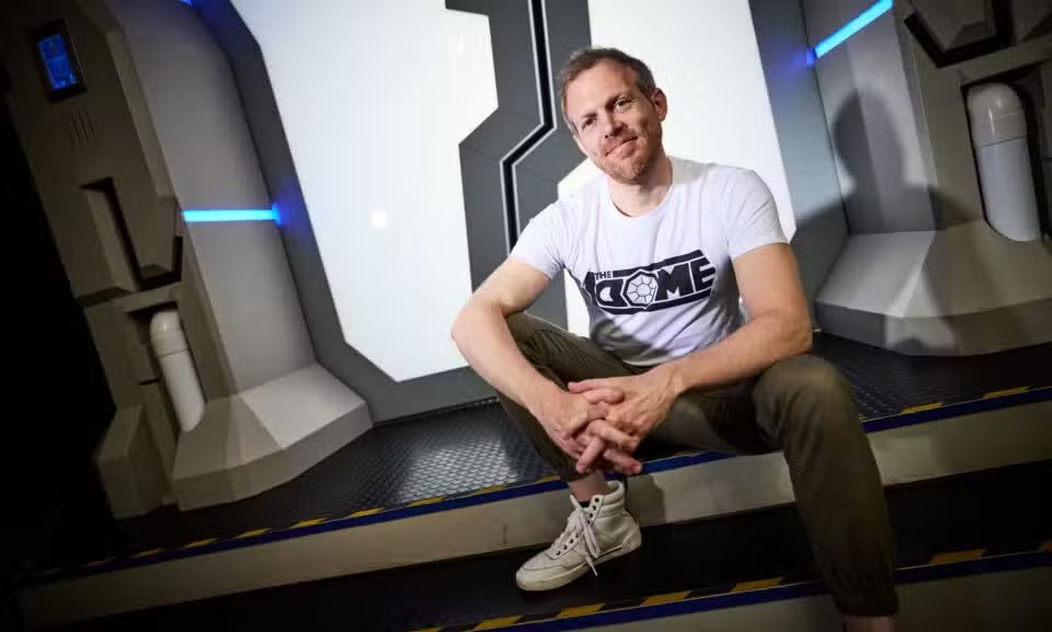 man with dark blond medium length hair and beard, sitting on some low steps in front of a futuristic doorway. He is wearing a white shirt with The Dome logo and olive green pants with white hightop shoes.