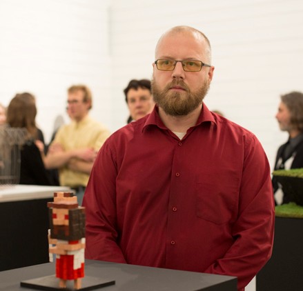 man with buzzed hair, glasses and full dark blond beard in red button down shirt sitting at a desk with a minecraft style figurine