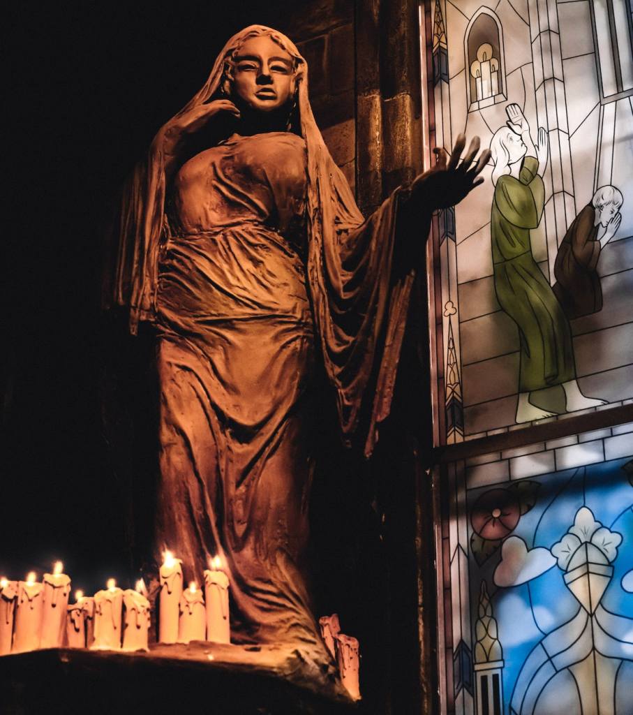 A statue of a woman holding her hand out beside stained glass panels. At her feet lay an assortment of candles, most of them are lit.