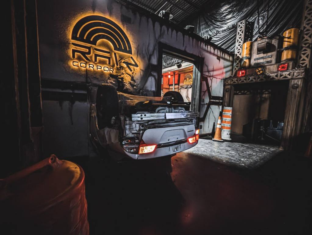 An upside down car crashed through a wall of a building bearing the RAIN Corporation logo.