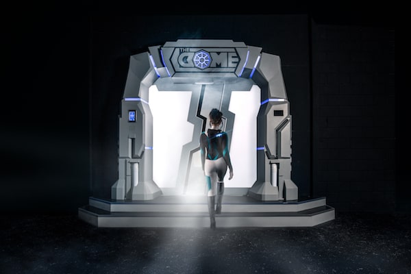In-game: A beautiful woman in a tight body suit approaching white and blue sci-fi entry way for The Dome.