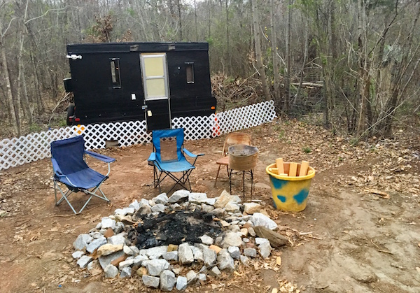 In-game: A trailer with a few folding chairs around an extinguished fire pit.
