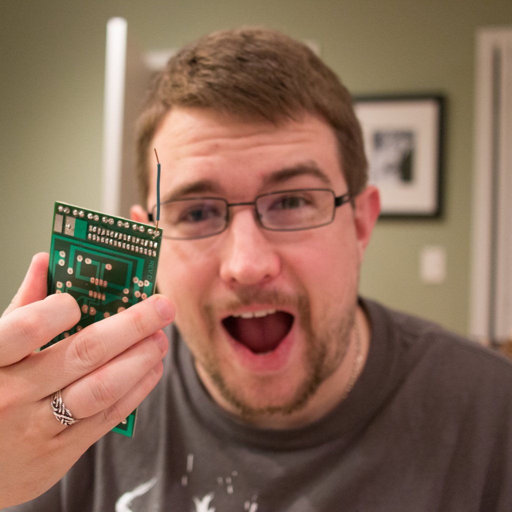 Man with rectangular glasses and short neat hair and beard looking gleefully at a circuit board he's holding in front of his face.