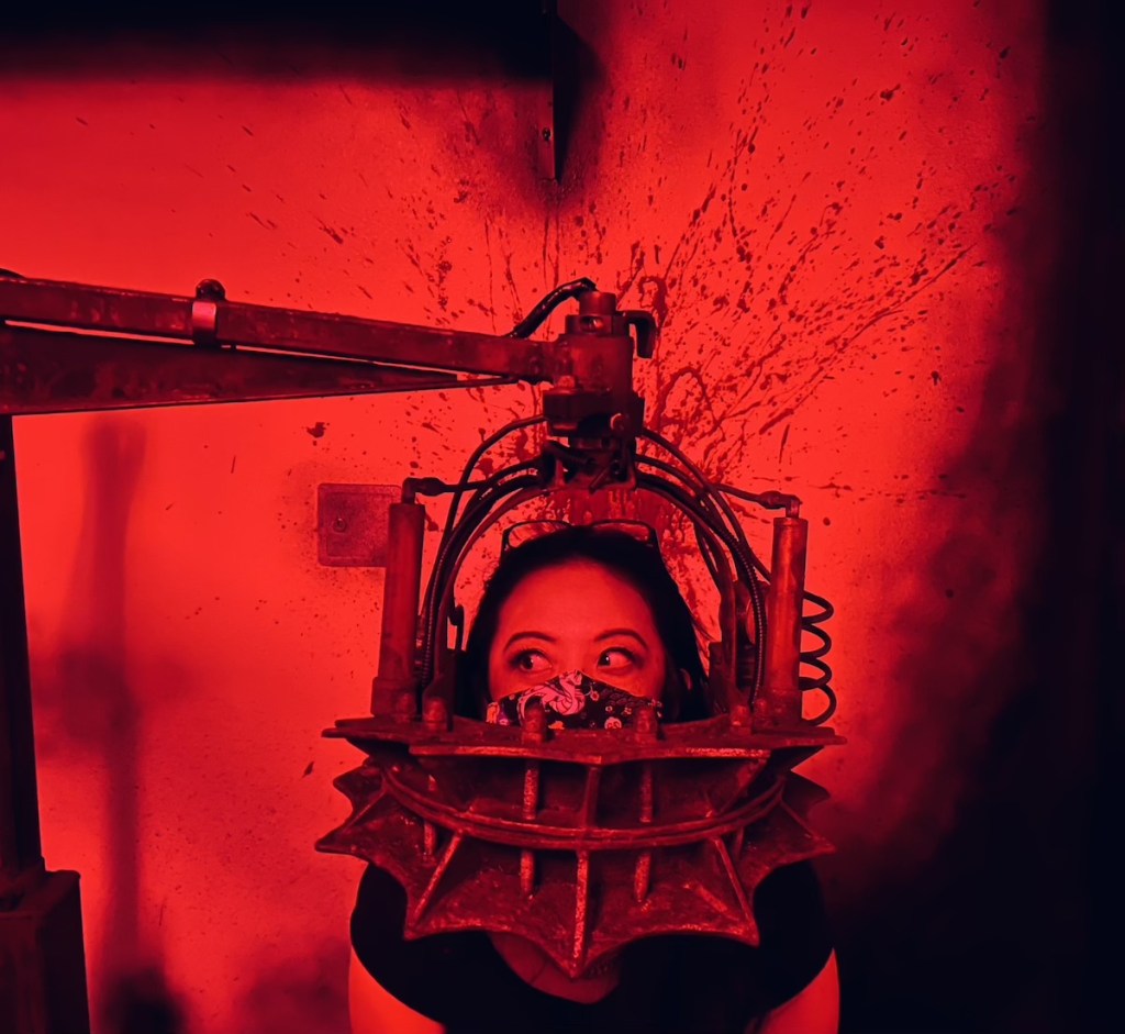 Peih-Gee Law with her head in an elaborate metal torture device. The wall behind her is blood spattered.