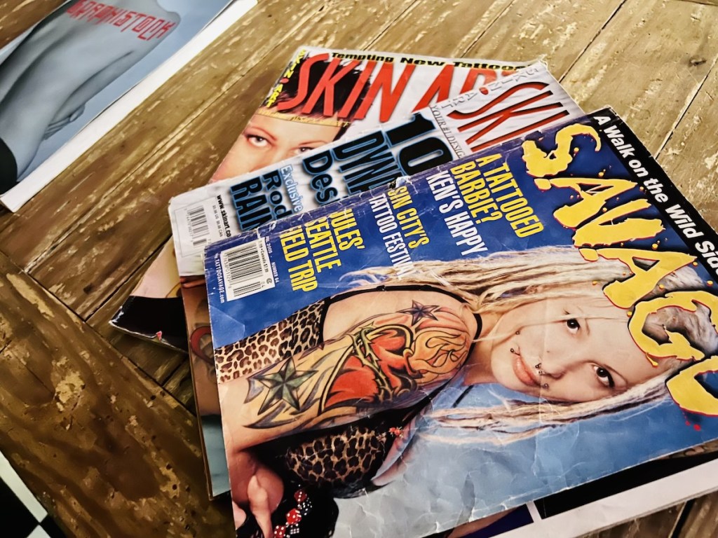 An assortment of tattoo magazines on a beat up wooden coffee table.