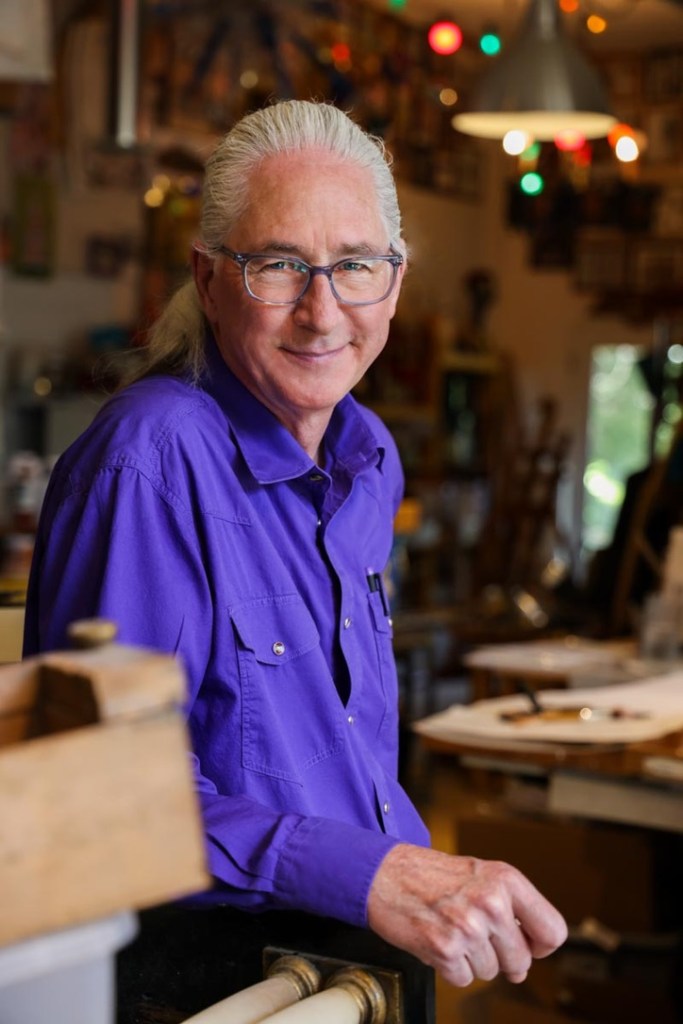 older man with long white hair in a low ponytail, smiling, wearing purple glasses and a long sleeved purple shirt, leaning on a counter in a workshop.
