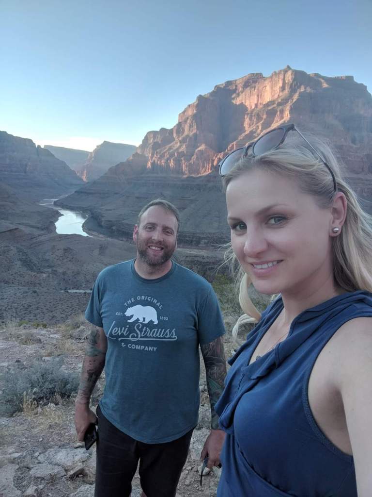 man and woman standing in wilderness. she is blonde, with sunglasses on her head and a ruffled blue tank top. He is bearded with short hair, blue t-shirt and tattoos on his arms.