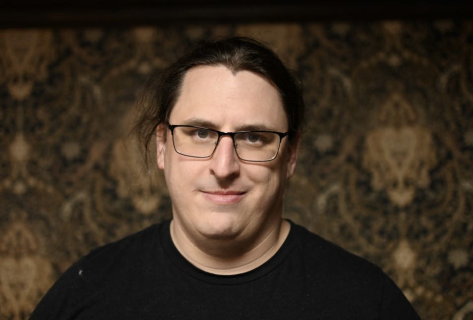 A clean shaven Man wearing a black t shirt and glasses with long hair pulled back into a ponytail with a few wisps escaping around his face.