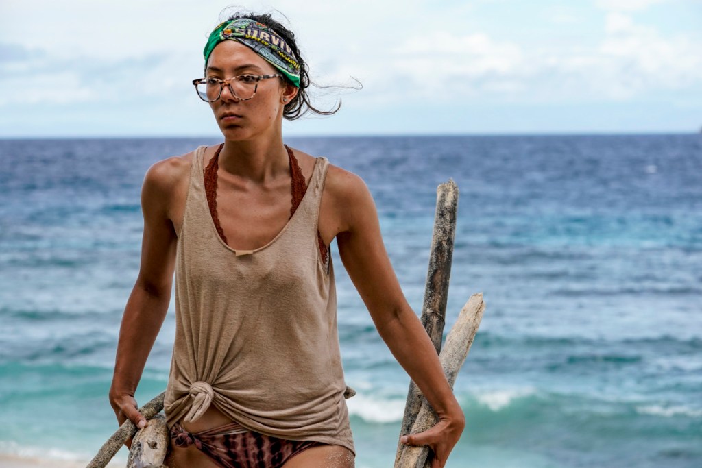 woman with surivivor buff on head wearing glasses, beige tank top and bikini bottoms hauling wood on a beach.