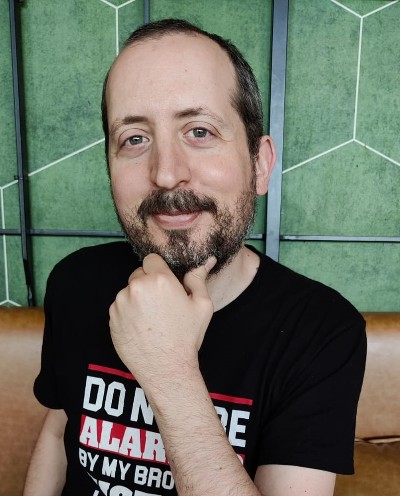 man smiling in short dark hair, short beard, holding his chin, wearing a black t-shirt in front of a green wall.