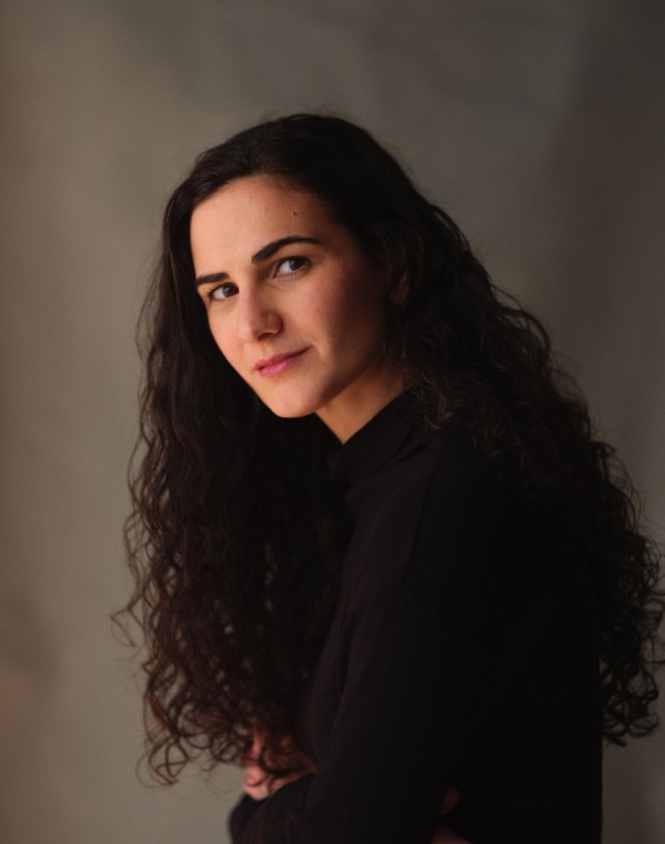 woman with long dark curly hair, strong brows and an enigmatic smile, wearing a black long sleeve mock turtleneck