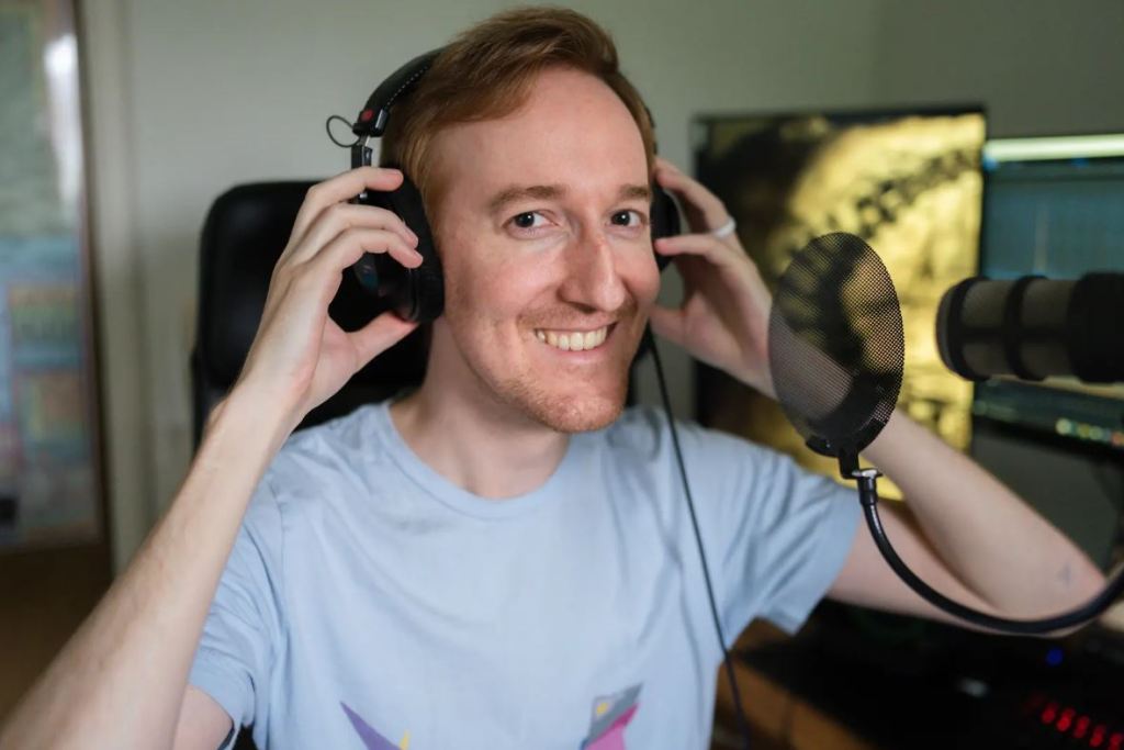 slender man with strawberry blonde hair sitting in front of microphone holding onto his headphones