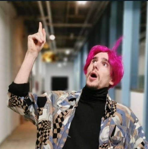 Man with pink hair and a brown goatee wearing a black turtleneck with a patterned jacket posing with one arm raised