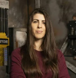 woman with long straight brown hair wearing a maroon color top inside an industrial looking escape room.