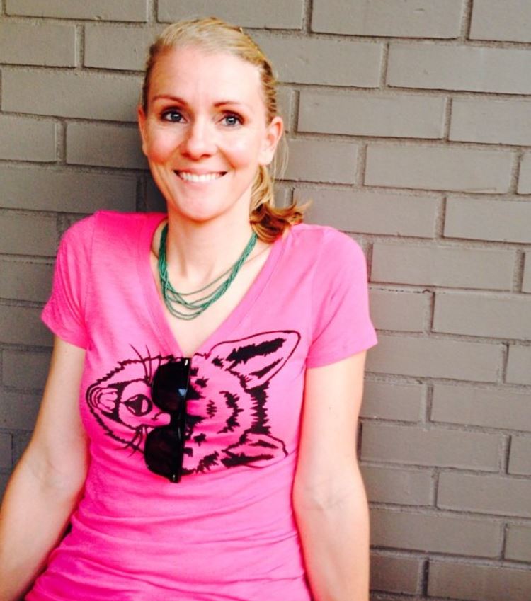 blonde woman smiling in pink t-shirt with a wolf face printed sideways. Sunglasses are hanging from the shirt collar covering the eyes of the wolf image.
