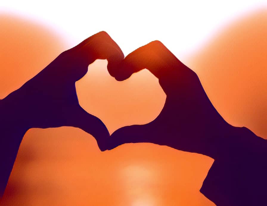 Hands of two people forming a heart with a sunset beyond.