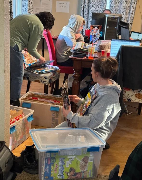 2 people sorting through flattened cereal boxes, surrounded by large plastic bins filled with flattened cereal boxes, and people on computers in the background