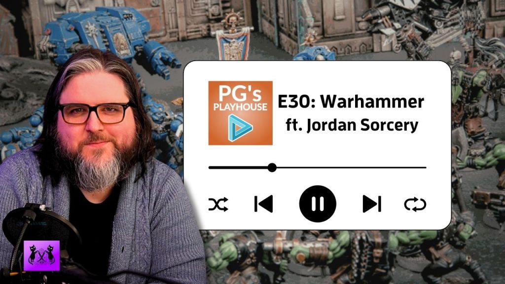 man with shoulder length brown hair with white streak at forehead, wearing glasses, with a medium length beard that's brown with a white center. He is wearing a grey cardigan. background of warhammer figurines.