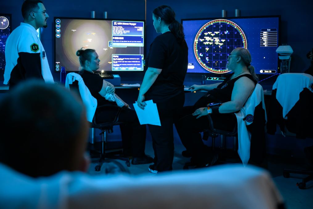 people standing and sitting in a headquarters style room with large monitors showing spacecraft information