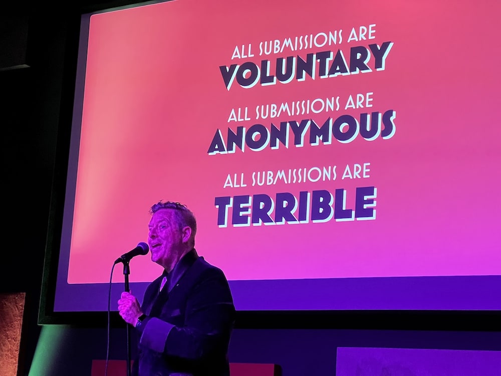 Brett Jackson in a spiffy jacket, MCing imagineer-do-wells. The projection behind him reads, "All submissions are voluntary, anonymous, and terrible."
