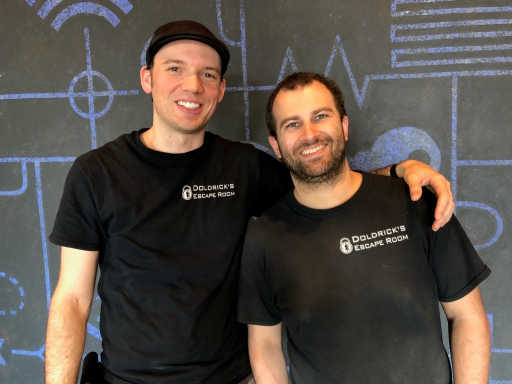 two men, both in black t-shirts that say "doldrick's escape Room". One is wearing a black newsboy had and is clean shaven. the other man is smiling with green eyes, short brown hair and a short beard.