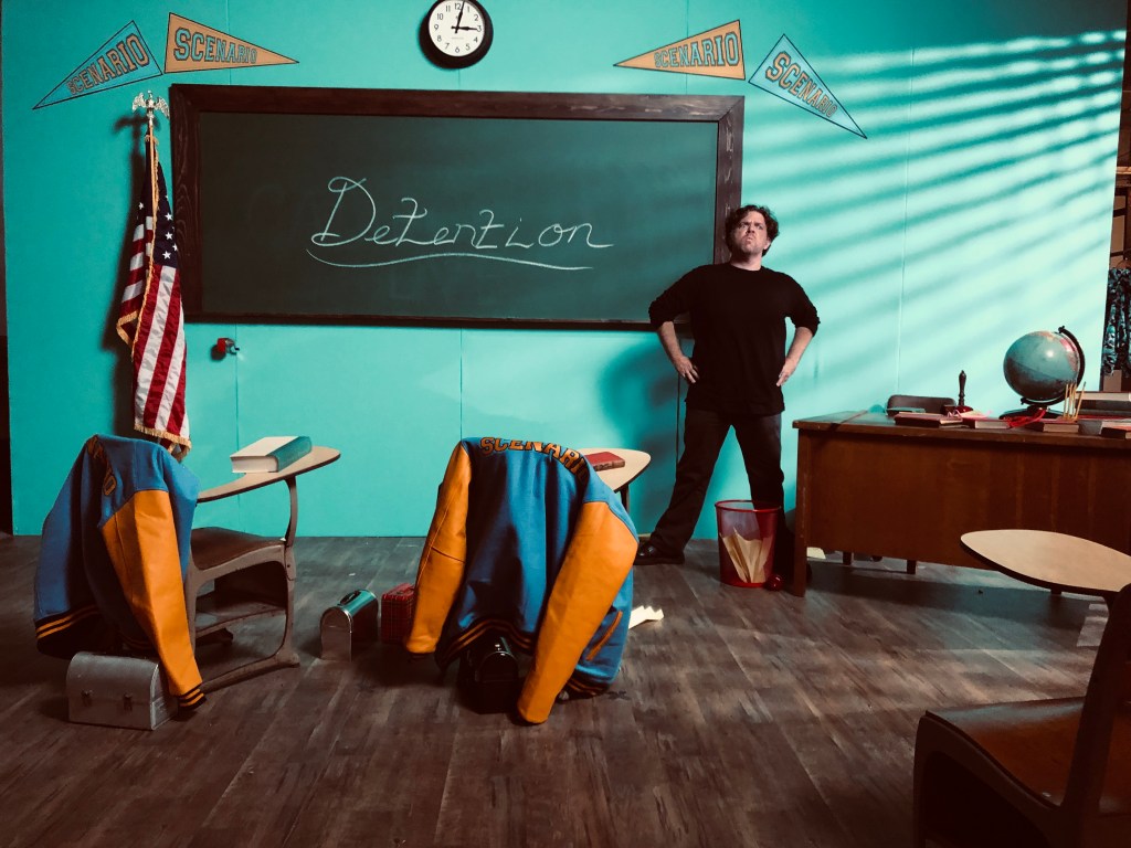 old fashioned looking classroom with teal walls. chalkboard says detention. there is a man wearing all black, standing with his hands on his hips and he is scowling.