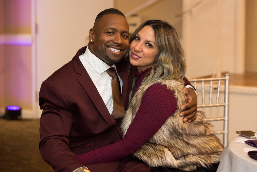 man in a plum color suit sitting and smiling while hugging a woman in a plum color dress with a furry vest