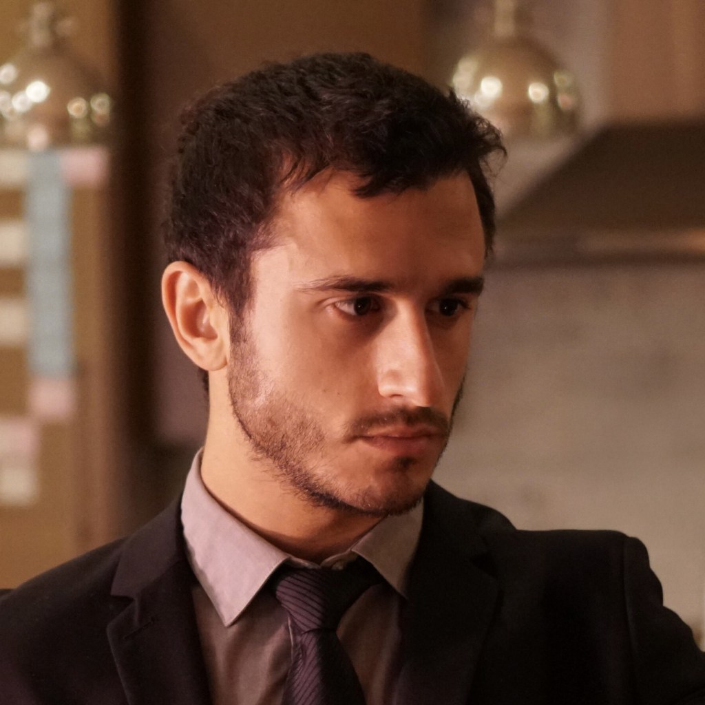 man with dark brown hair, short beard, in a dark suit and tie. he is looking to the side with a serious expression