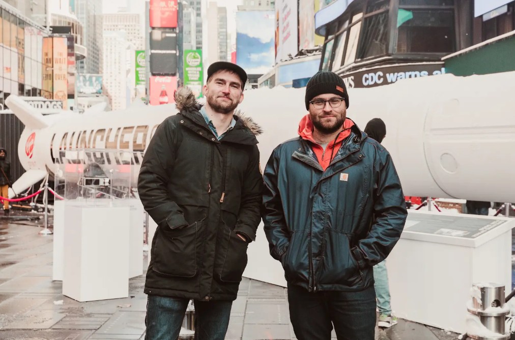 Two men with beards and jackets in jeans, standing in Times Square in NYC in front of a Virgin Rocket launcher model