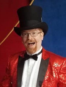 man with ginger red hair, curly mustache and beard with glasses wearing a black top hat and a red sequined tuxedo. He is smiling open mouthed in surprise.