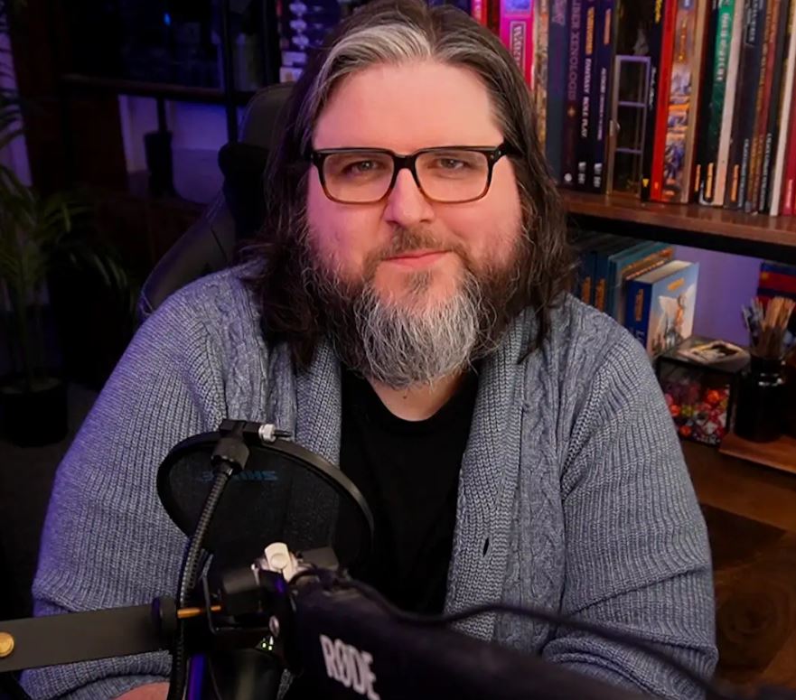 man with shoulder length brown hair with white streak at forehead, wearing glasses, with a medium length beard that's brown with a white center. He is wearing a grey cardigan, sitting in front of a bookshelf with a mic setup in front of him.
